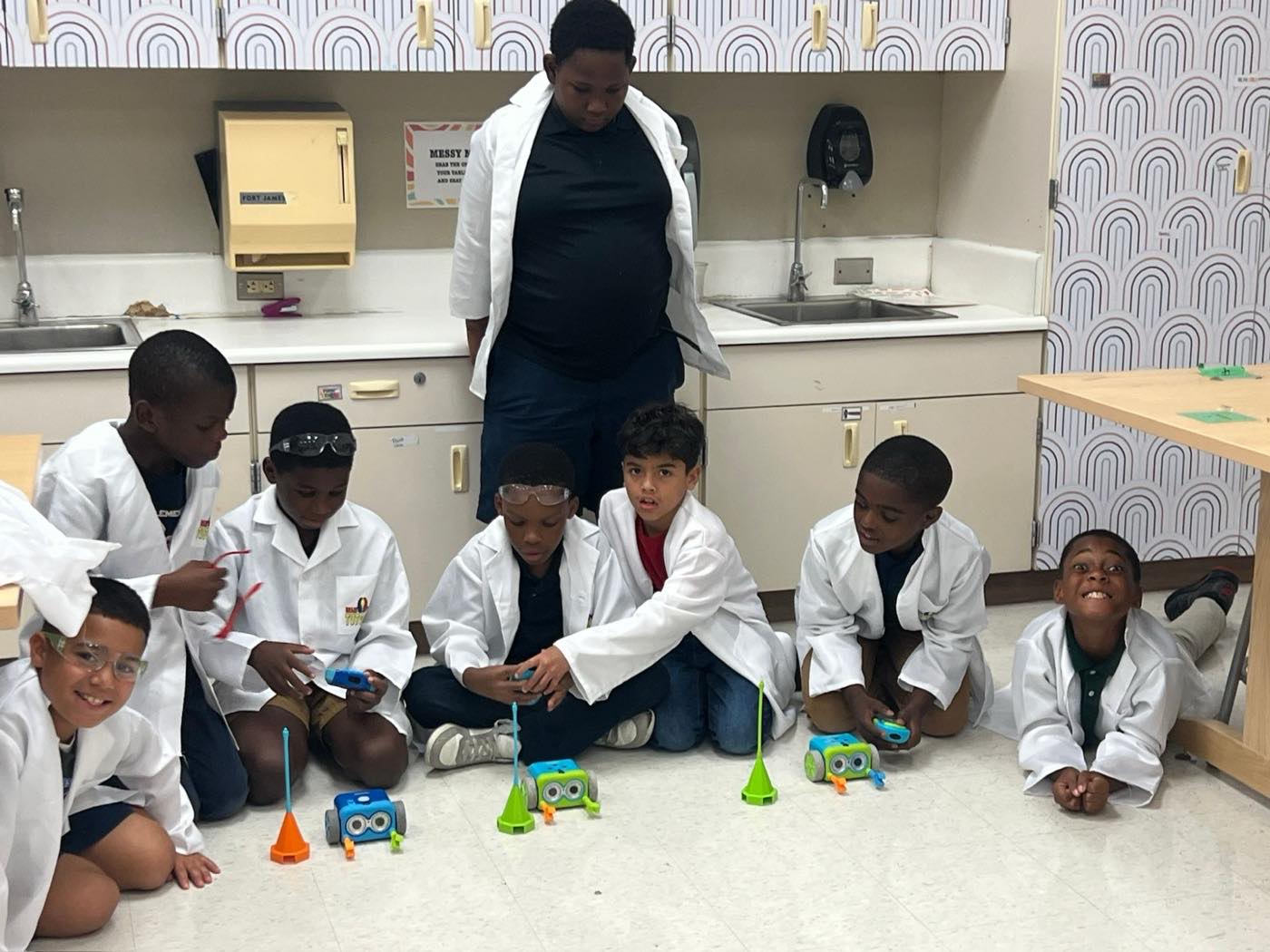 A class of students in BrainBuzzed lab coats running coding robots on the floor